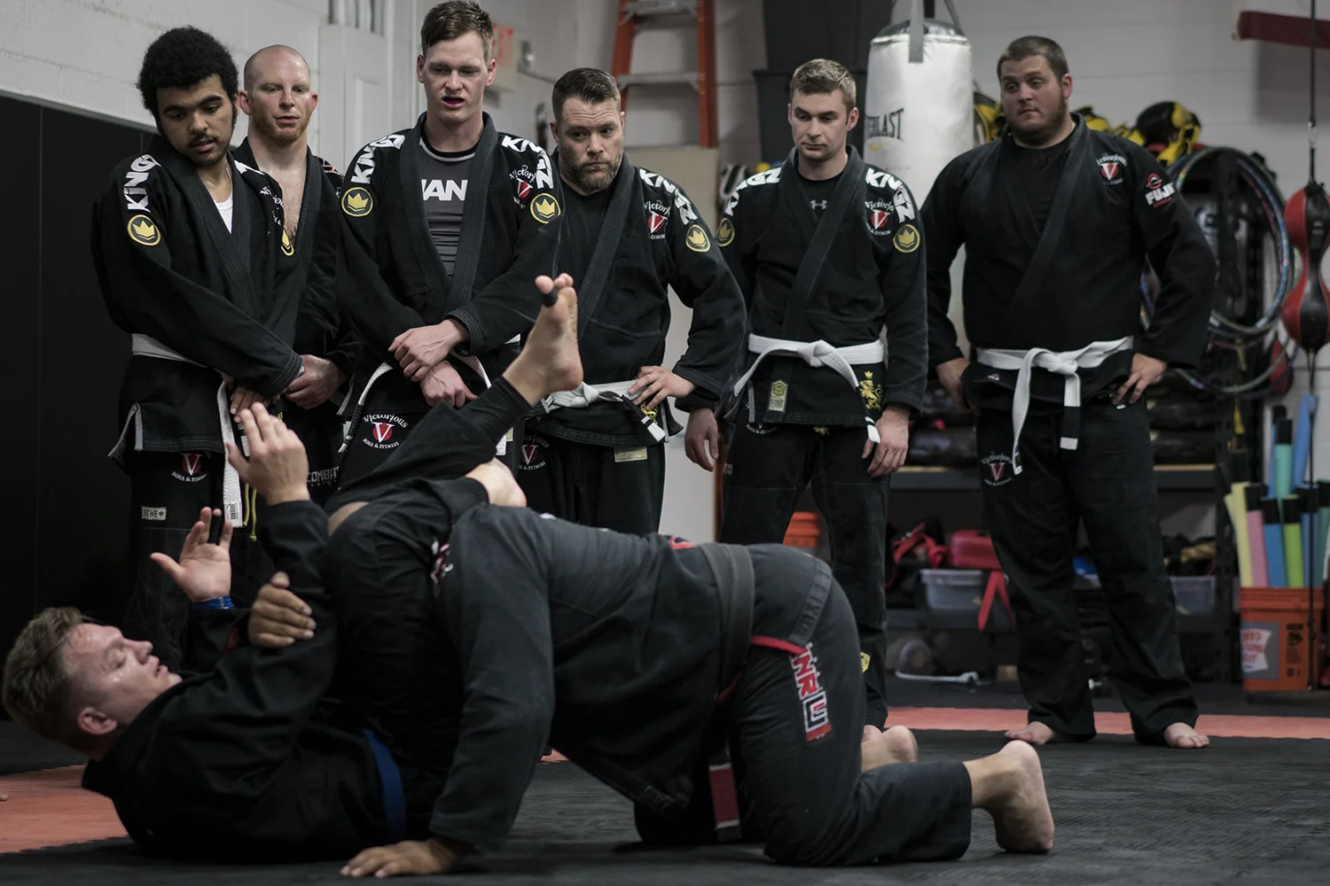 A row of students in dark gis standing and watching two people demonstrate a technique on the mat.