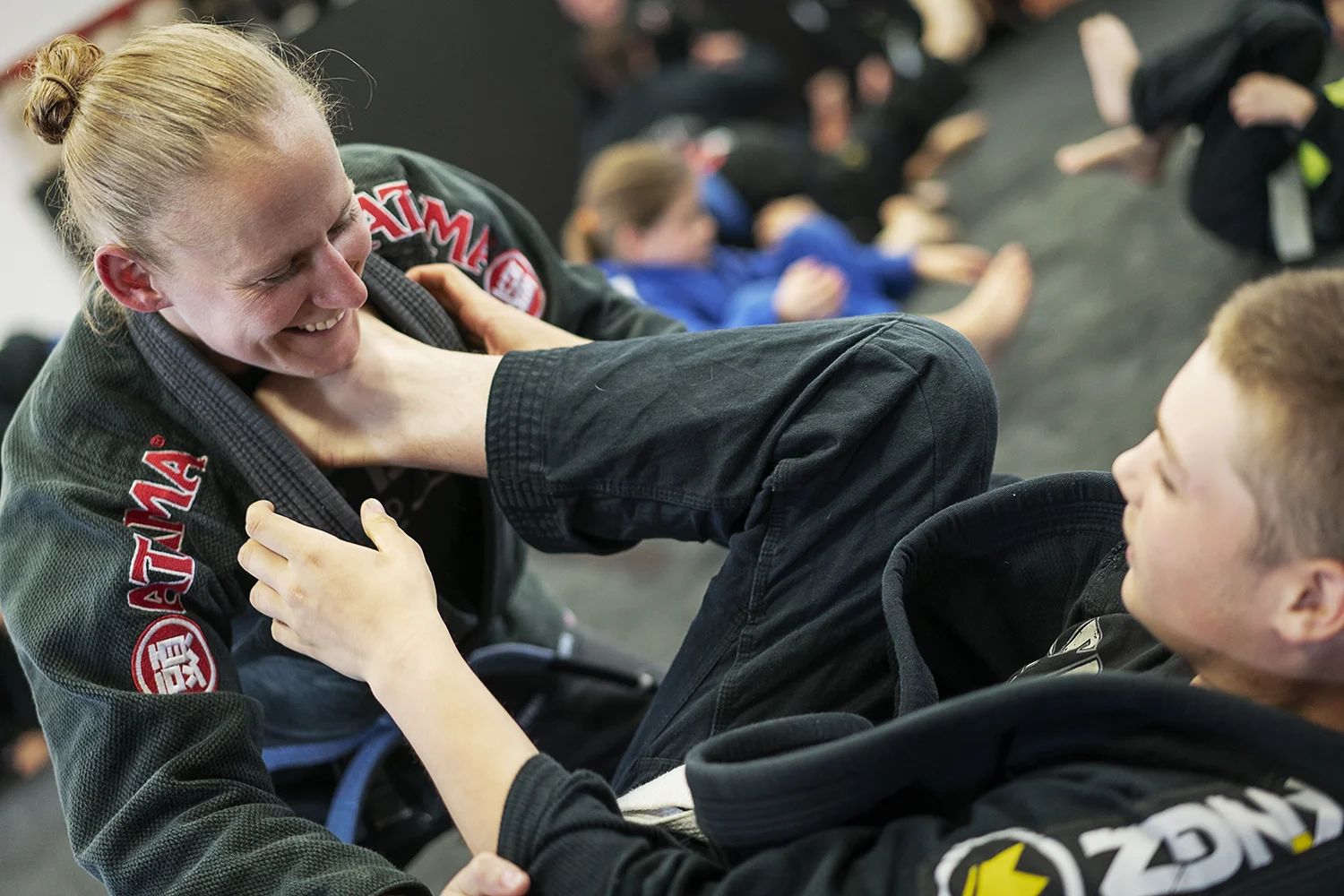 A coach in a dark gi guiding a young student through a jiu-jitsu position, with other children training behind them.